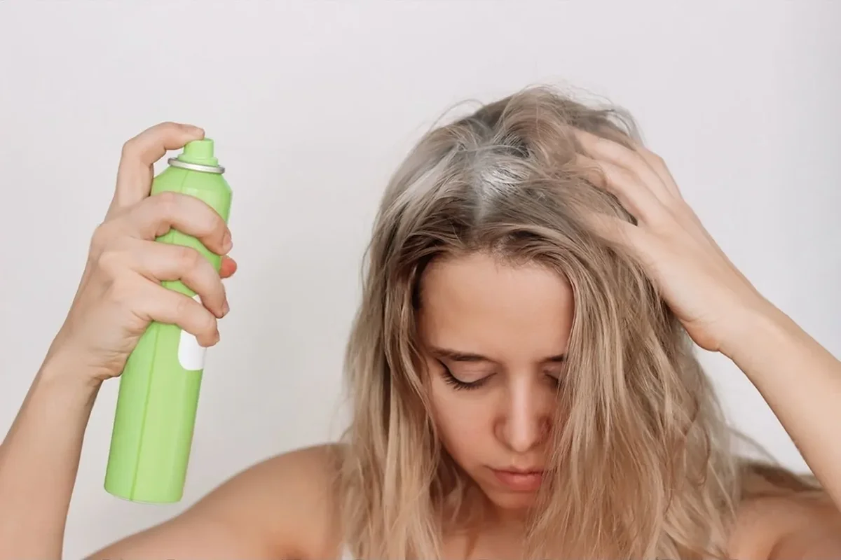 Person applying dry shampoo to blonde hair with a green spray bottle.